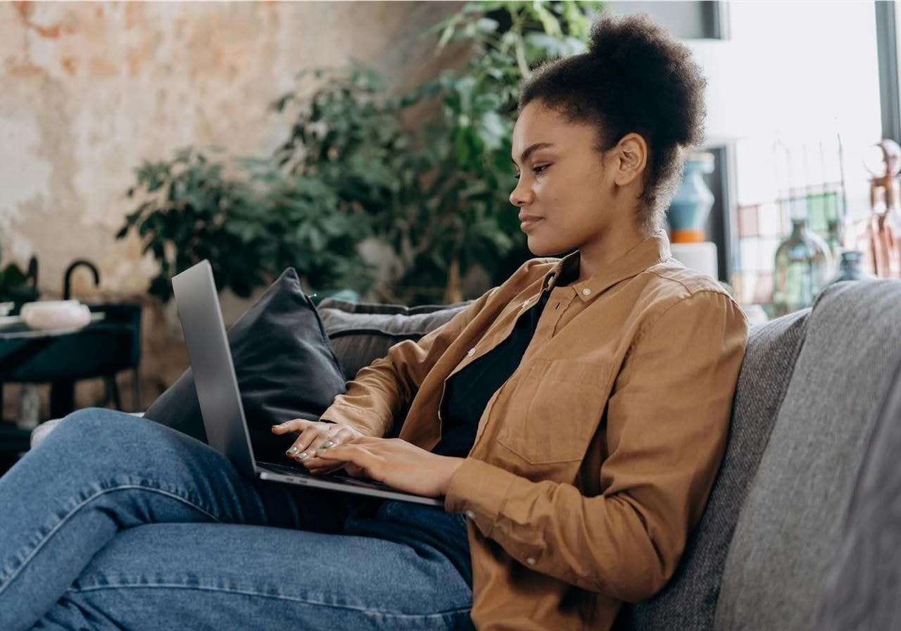 A young woman sitting on a grey couch with a laptop on her lap.